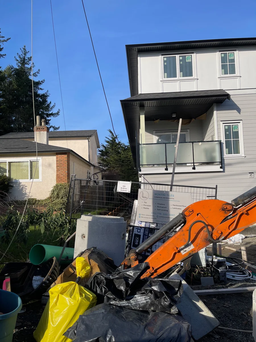 Construction site at 990 McBriar Avenue: a new three-storey multiplex behind site fencing, with an excavator arm, stacked building materials and bagged debris in the foreground, next to an older single-storey house.