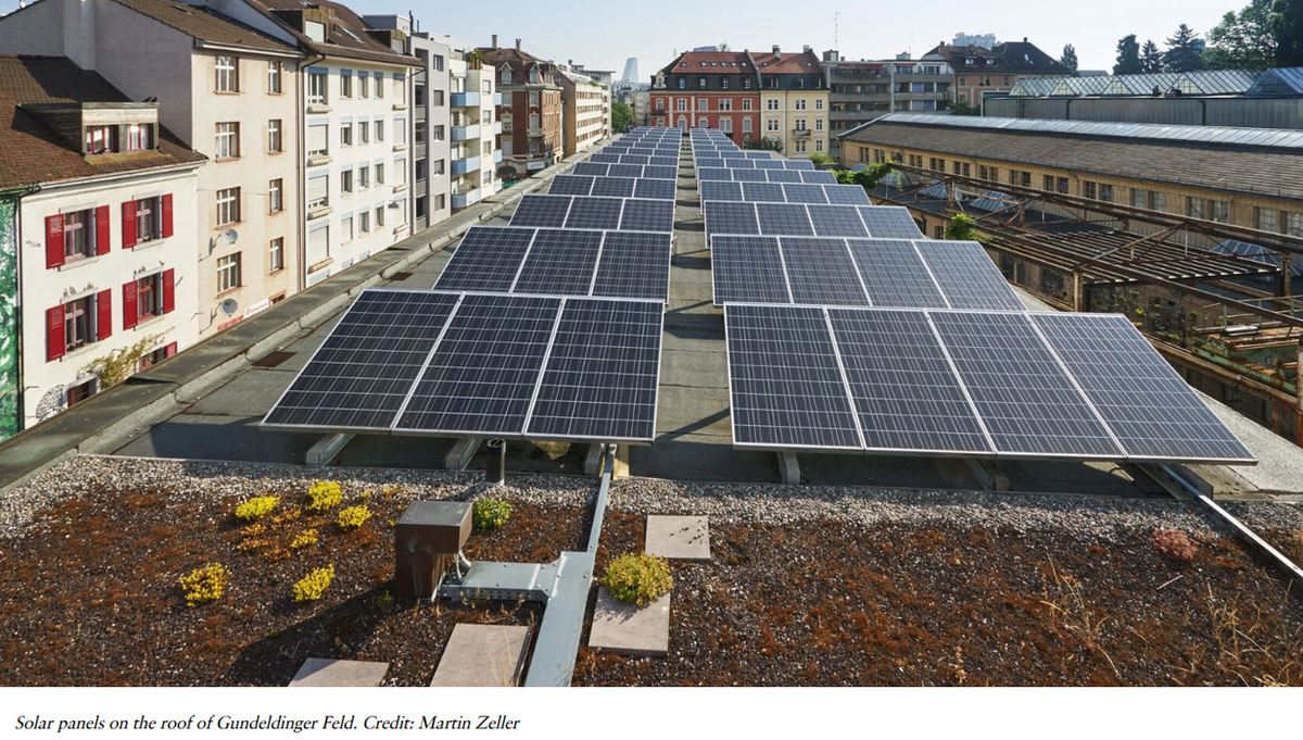 Rows of solar panels mounted on the green roof of Gundeldinger Feld, with Basel apartment buildings and rooftops behind.