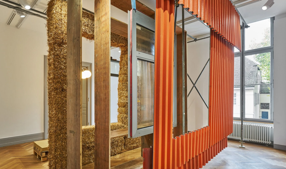 An exhibition mock-up of reused construction: straw-bale insulation between salvaged timber posts, a second-hand window in its frame, and a hanging panel of orange corrugated sheet.