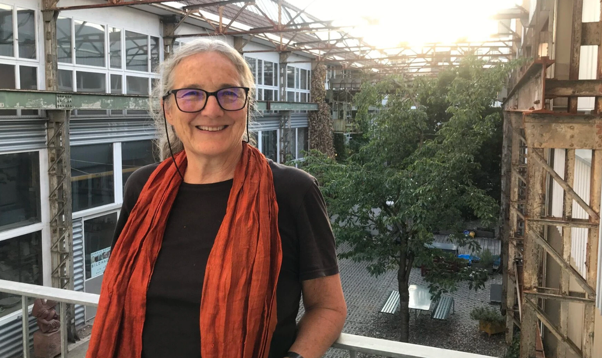 Swiss architect Barbara Buser, smiling, with grey hair, dark glasses and an orange scarf, standing on a walkway above a converted factory courtyard.
