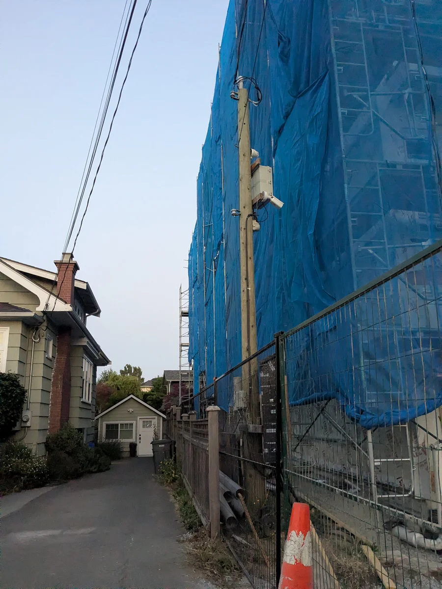 A narrow residential lane. On the left an older house with a brick chimney and a small garage behind it. On the right a construction site wrapped in blue tarpaulin and scaffolding, built out to the fence line, with a utility pole, stacked pipe and a traffic cone in the strip of ground beside it.