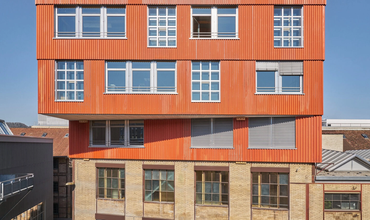 The K.118 building in Winterthur: three upper storeys clad in bright orange corrugated metal, cantilevered over the yellow brick ground floor of the older factory they were built onto.
