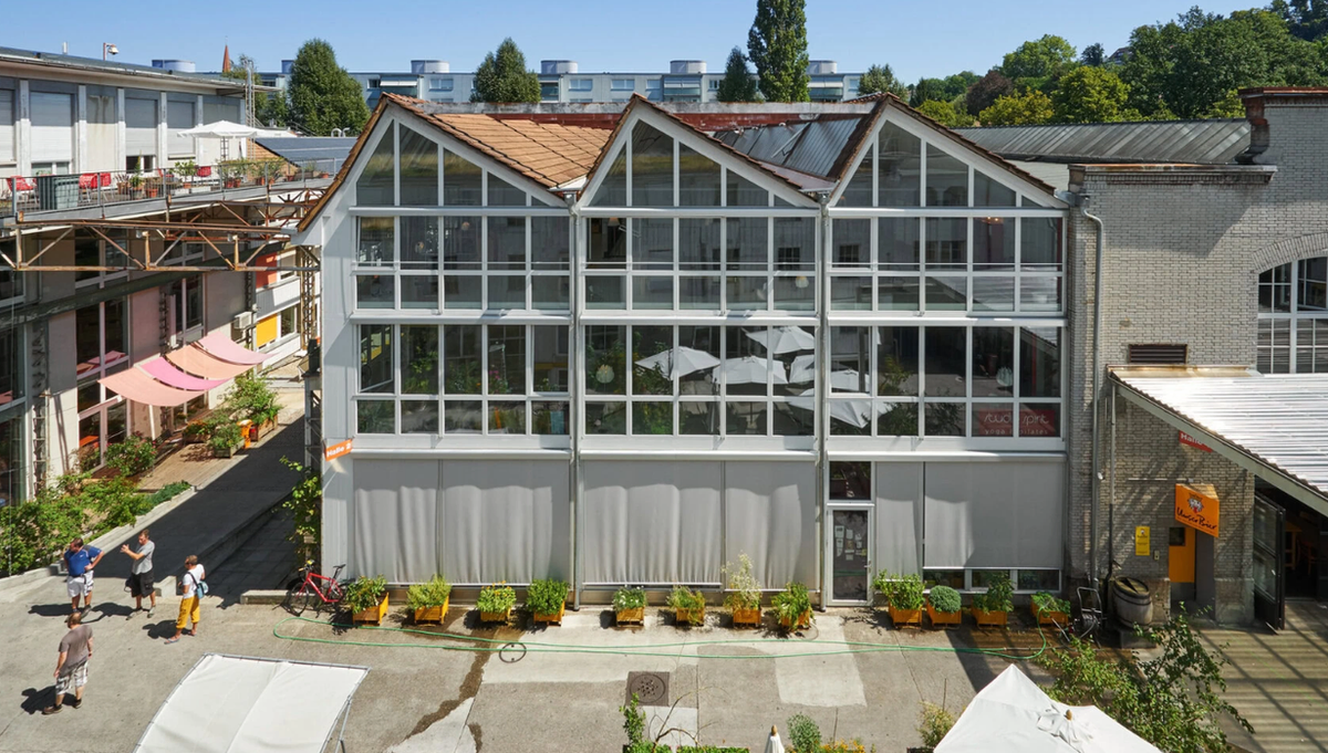 A tall glazed hall with a gabled steel-framed facade in a converted factory courtyard, planters and folded parasols along its front and people walking past.