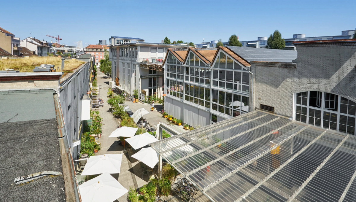 Gundeldinger Feld seen from a rooftop: a glazed hall, café parasols, planted terraces, green roofs and solar panels across the converted factory complex.