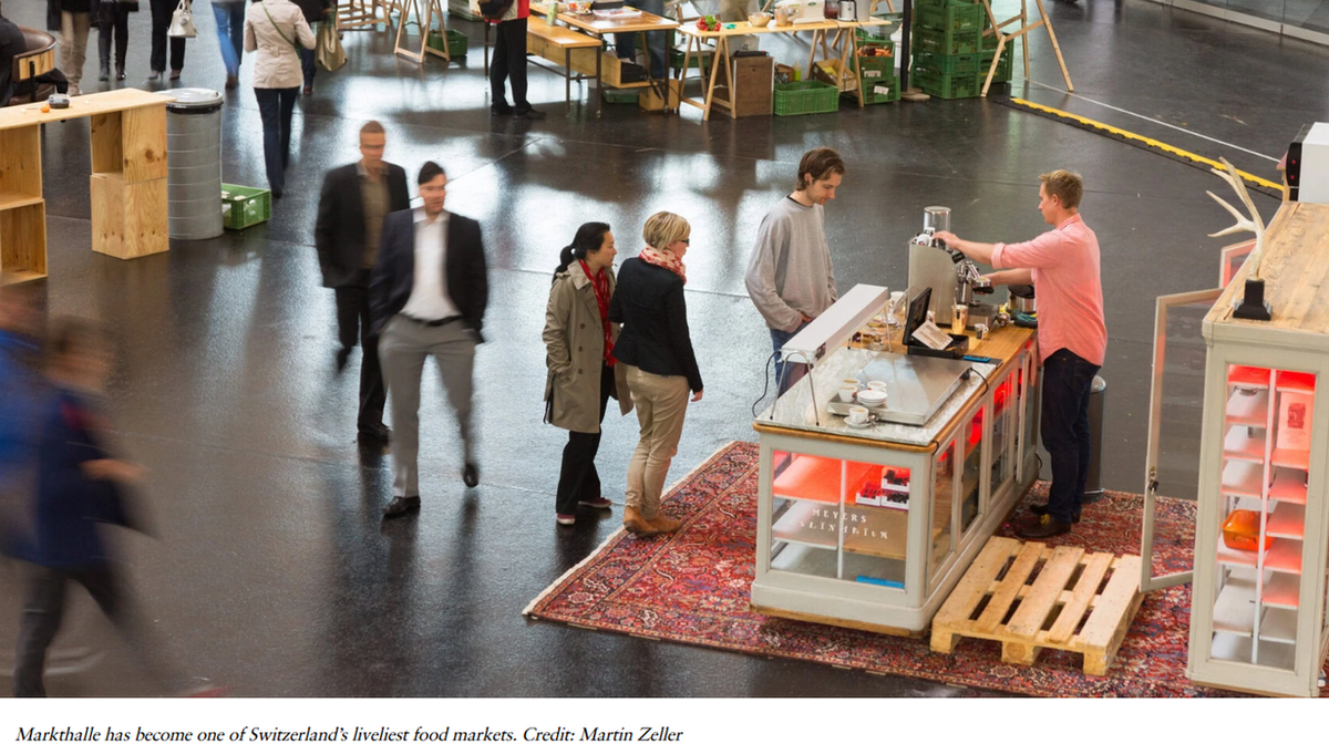A coffee stall standing on a patterned rug inside the converted Markthalle, with people queuing at the counter and others walking past.