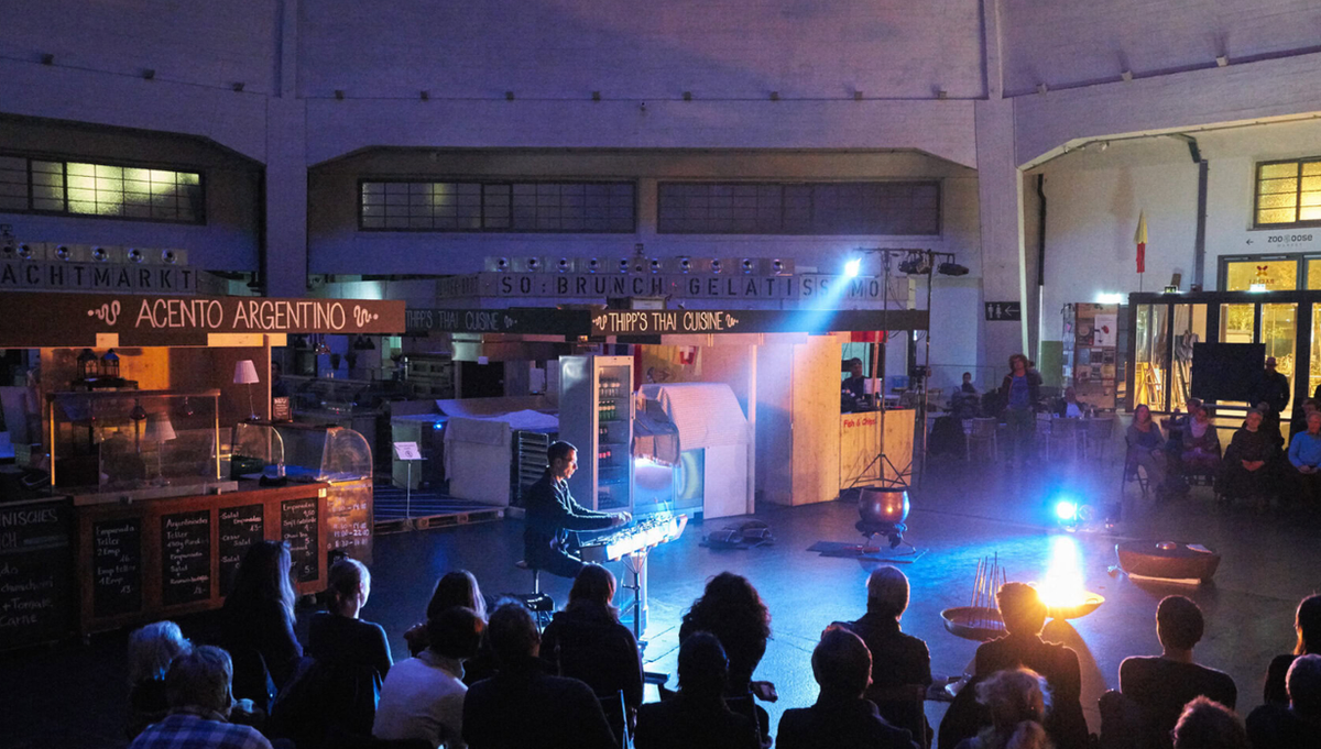 The domed Markthalle in Basel at night, food stalls ringing the hall and an audience seated on the floor watching a musician perform under a spotlight.