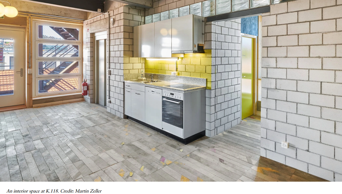 An apartment interior at K.118: a compact kitchen against white-painted blockwork, with reclaimed parquet flooring in mixed tones and a salvaged window beside the door.