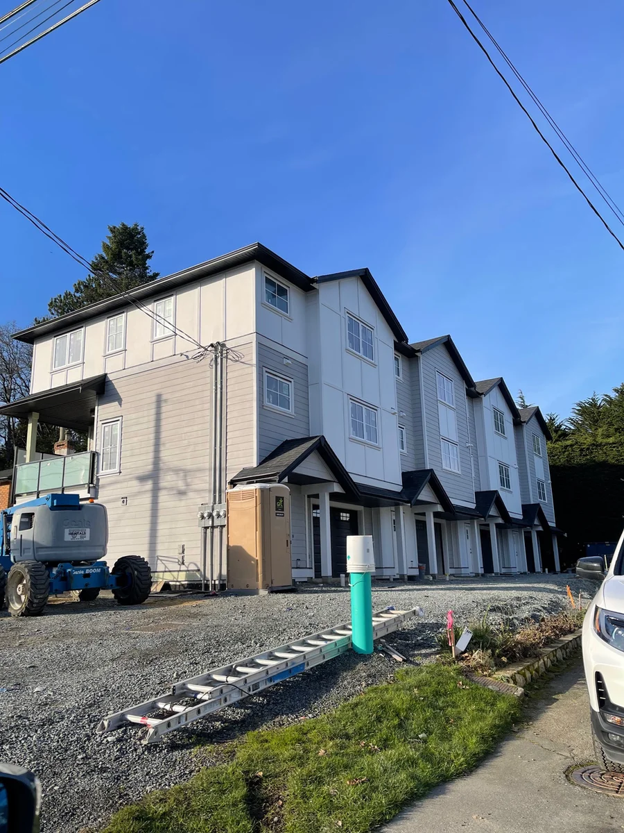 A row of newly built three-storey townhouses on a gravel lot, still under construction, with a boom lift, a portable toilet and a ladder on site.