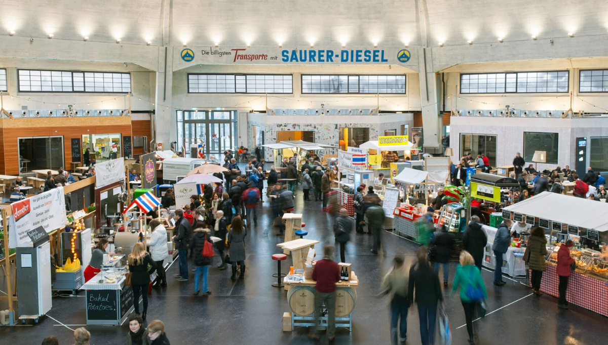 The interior of the Markthalle busy with shoppers among food stalls, under a preserved painted sign reading “Die billigsten Transporte mit Saurer-Diesel”.