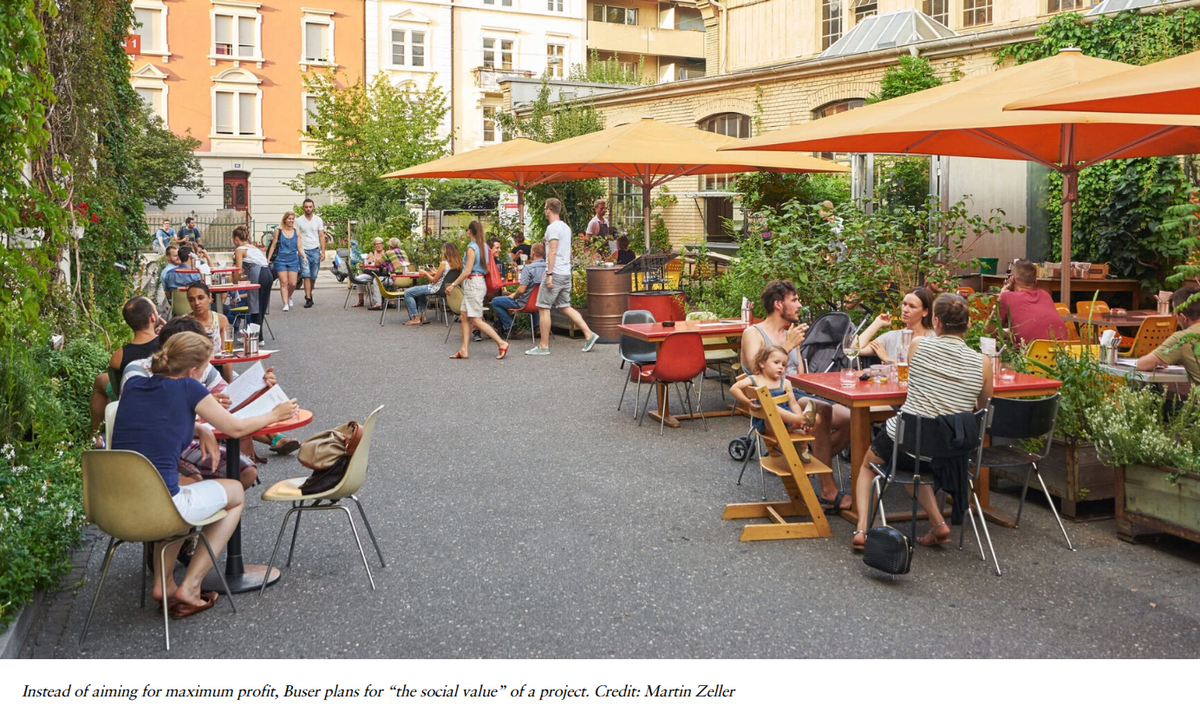 People eating and drinking at tables under orange umbrellas in a converted industrial courtyard planted with trees and climbing greenery.