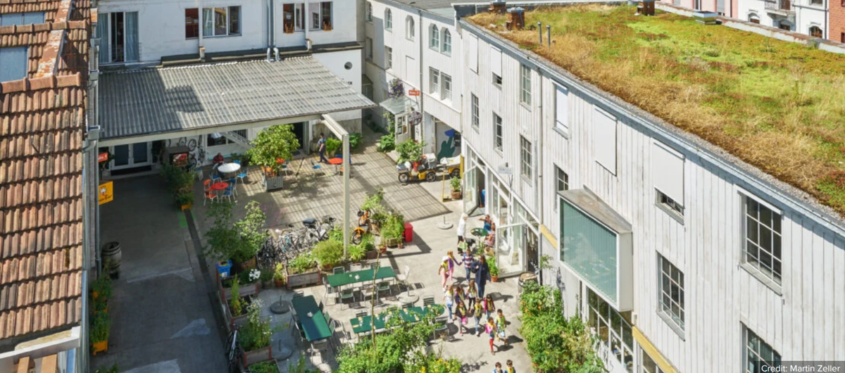 Aerial view of the Gundeldinger Feld courtyard in Basel, a converted factory with a green roof, planters, café tables and children walking between the buildings.
