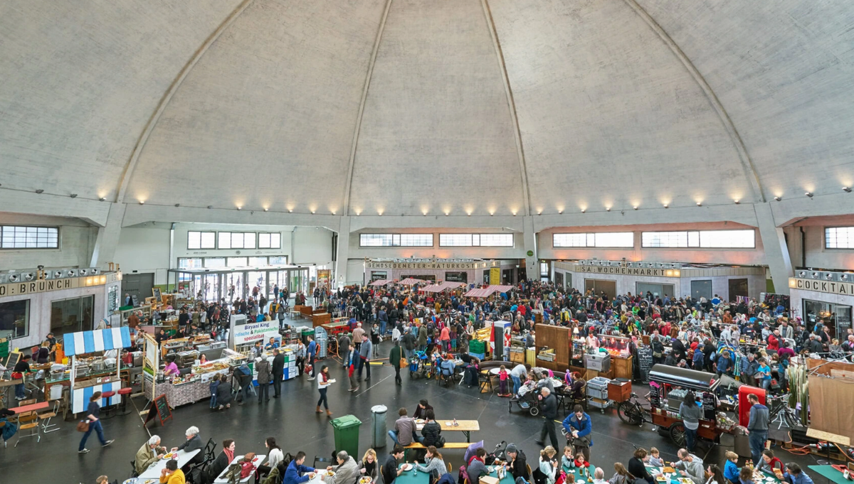 The concrete dome of the Basel Markthalle seen from a balcony, with market stalls around the edge and crowds eating at long tables below.