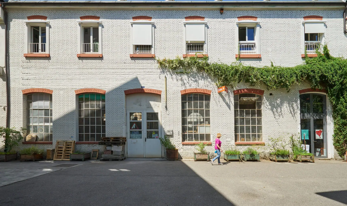 The white-painted brick facade of a converted factory building, with tall arched windows, climbing greenery along the upper floor and planters at ground level.