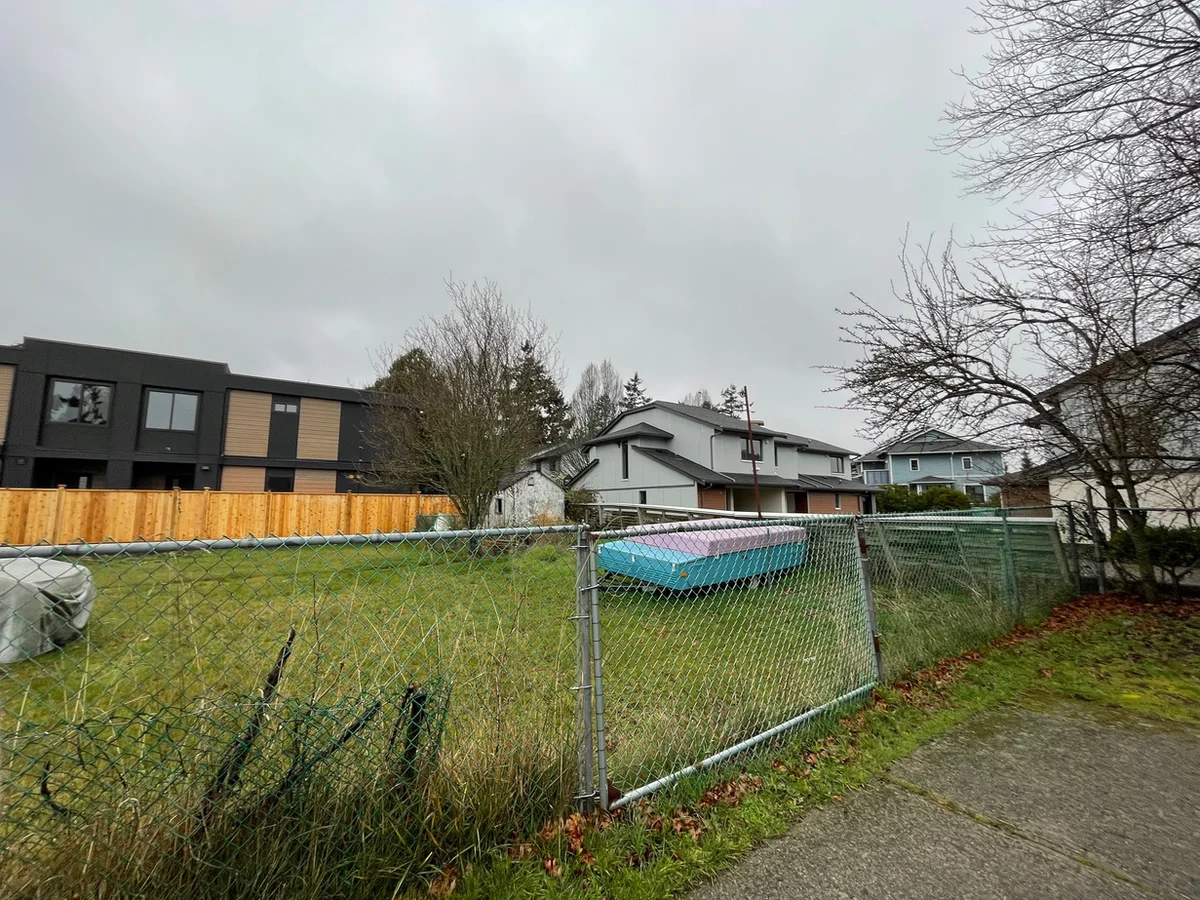 A fenced vacant lot of long grass with discarded mattresses piled at the back, a new dark grey multiplex on one side and older houses beyond.