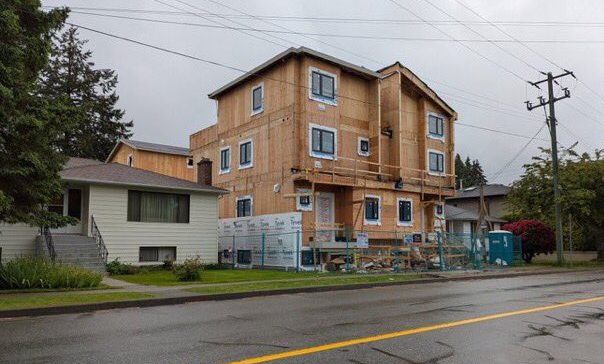 A four-storey wood-frame building under construction on a residential street, rising directly beside a single-storey house.
