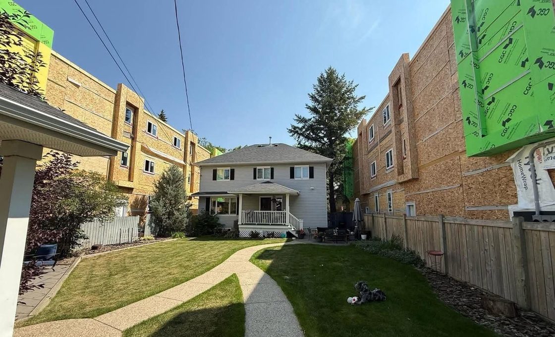 A two-storey house with a porch and lawn, hemmed in on both sides by three-storey blocks still in bare sheathing and housewrap, both built out to the property line.