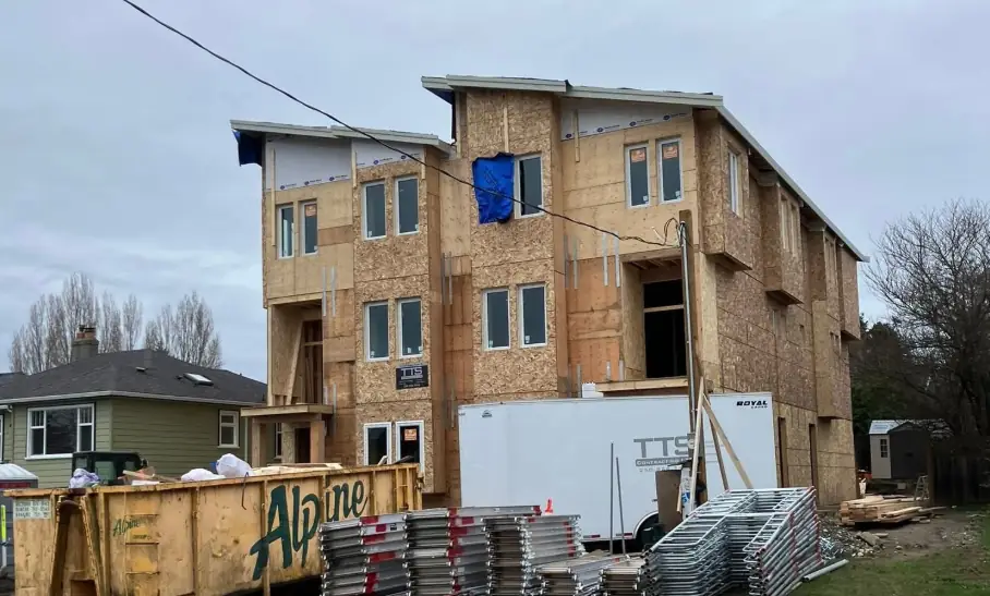 A three-storey building under construction in bare wood sheathing, with a skip and stacked scaffolding in front of it, standing directly beside a small single-storey green house.