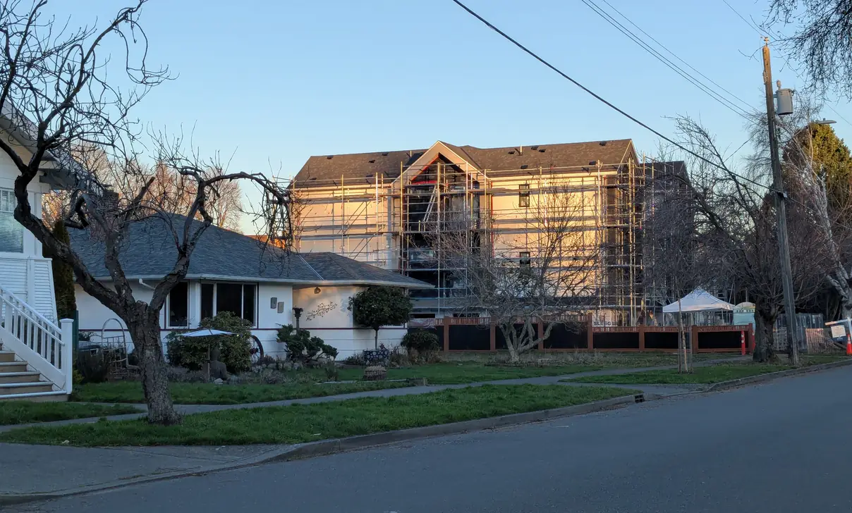 A tall wood-frame building in scaffolding filling the width of a residential lot in Victoria, rising several storeys above the single-storey bungalow and garden in front of it.