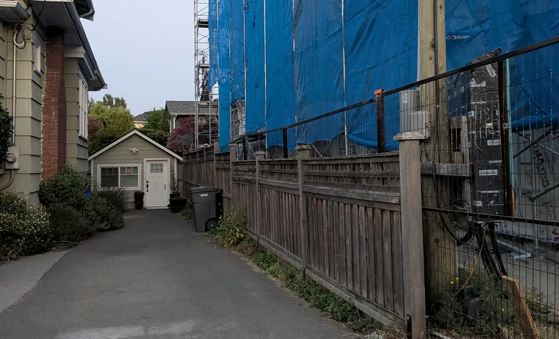 The same Victoria lane looking further down it: the blue-shrouded building running the whole length of the right-hand side above a wooden fence, with the neighbouring house and its garage small on the left.