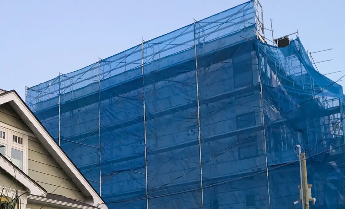 A building wrapped in blue mesh and scaffolding filling almost the whole frame against a clear sky, with the roof gable and upstairs window of a neighbouring house occupying only the lower left corner.