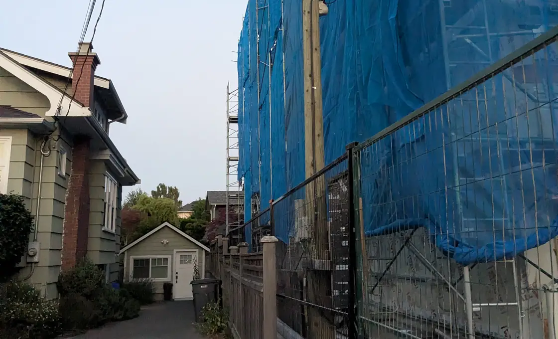 A Victoria lane. On the left a character house and its single-storey garage; on the right a building several times their height, wrapped in scaffolding and blue mesh, running the length of the lot to the fence line.